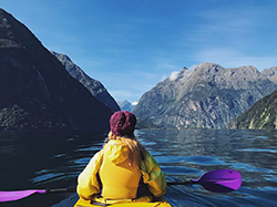 A young person wearing a bright yellow jacket in a boat in the foreground, facing a vast scenery of mountains and blue sky.