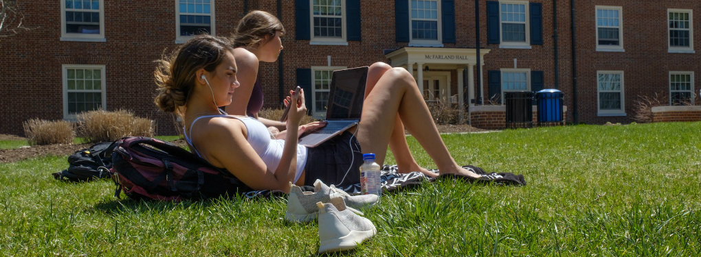 2 students recline on the ground near McFarland Hall. One looks at her phone and the other uses a laptop computer