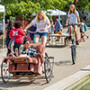 A girl rides in a recliner chair that is attached to a bike driven by a female student while another female student rides a very tall bike in the background
