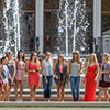 A group of females singing in front of a fountain shooting water up into the air