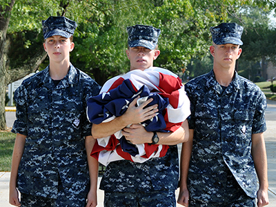 3 NROTC students with the middle student holding a wadded up flag in his arms.