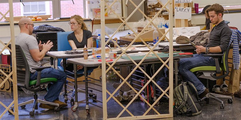 Professor and 2 students converse at a rectangular table in the studio, as seen through a lattice