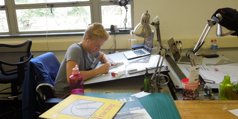 Seen from a distance, a student works at a drafting desk in a studio, surrounded by drawing supplies and a laptop comptuer