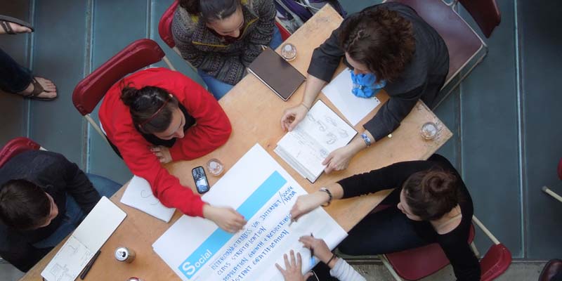Seen from overhead, several students seated at a rectangular table collaborate on a project