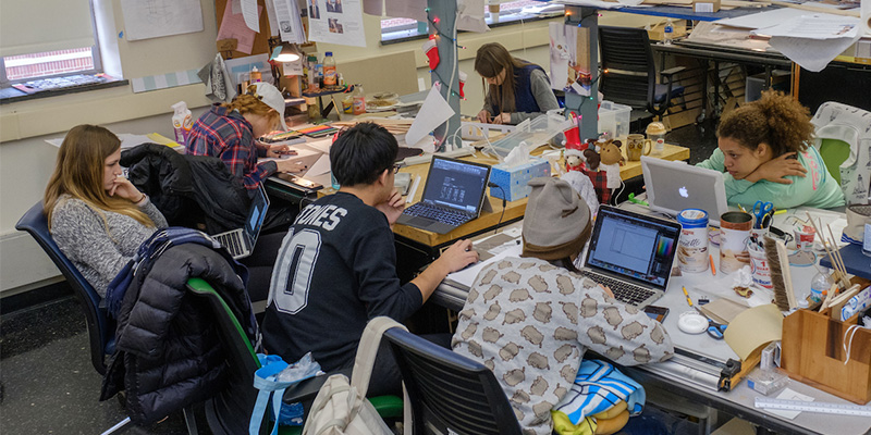 A group of students seated at rectangular studio tables work on projects using art supplies and computers. The tables are covered with assorted supplies such as drawing and cutting implements