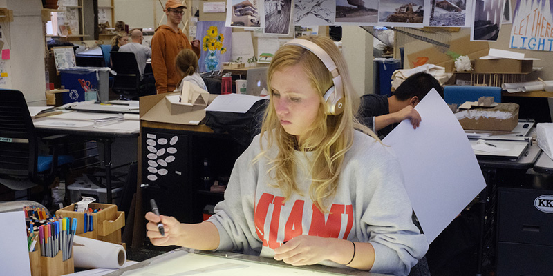 A woman wearing a Miami shirt and headphones is seated at a worktable. She holds a pen and works on a drawing