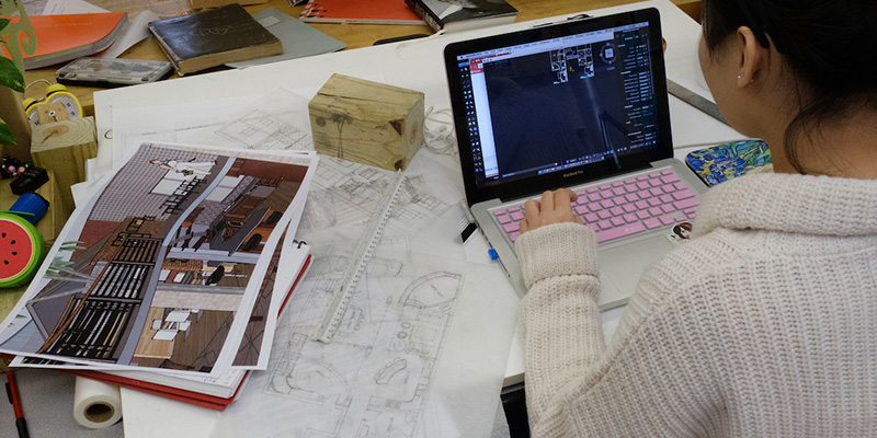 Viewed from behind the shoulder, a woman works on a computer. Architectural drawings and sketches are spread out around her.