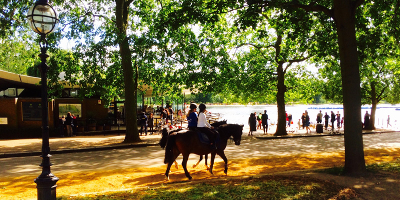 People in the foreground ride horses while others gather at the water's edge in Hyde Park
