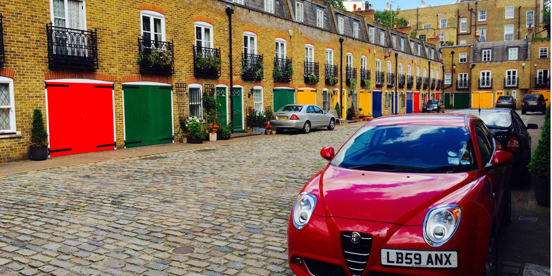 London Mews. A cobblestone street is lined with colorful buildings. Cars are parked along the street