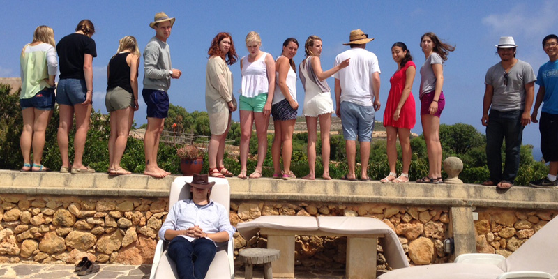 Students stand on a stone wall as a man wearing a cowboy hat over his eyes reclines in a lounger in Malta