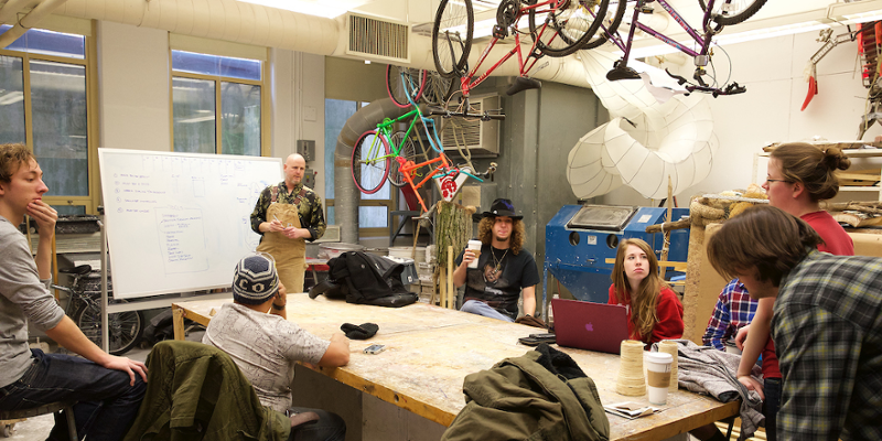 A professor stands at a chalkboard in the sculpture studio as students around a worktable participate in class.