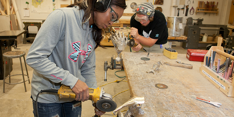 A student wearing ear and eye protection works with power tool to shape a metallic piece