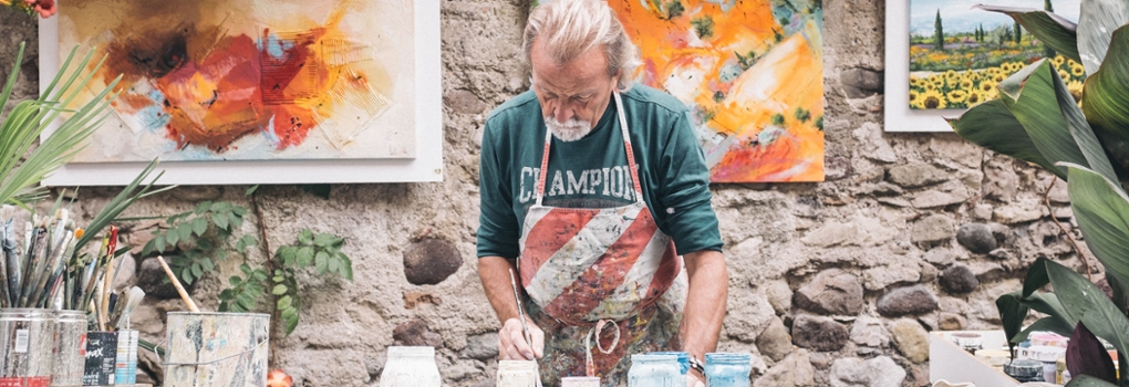 A painter stands at his bench with his supplies spread out in front of him and some completed works on the wall behind him