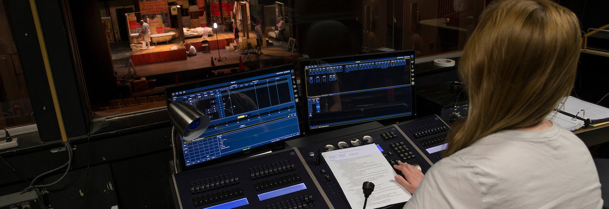 A student operates a theatre lightboard in foreground, with a view of the illuminated stage