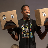 A theatre student performs as he holds two box-shaped puppets