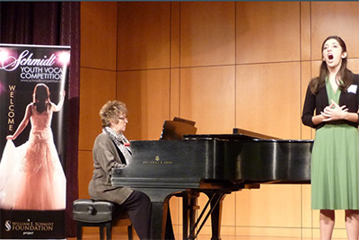 A student sings onstage as a pianist accompanies her. Schmidt Vocal Institute Banner is visible at left.
