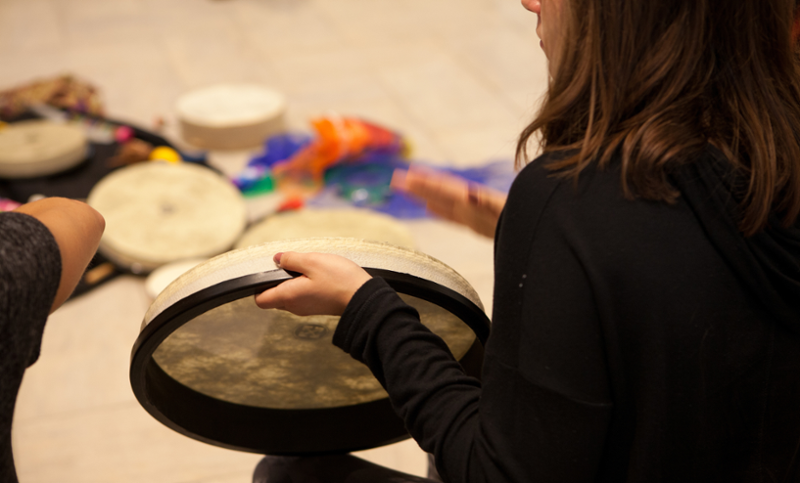 Hand drums are arranged on the classroom floor in the background. Seen from behind, a student plays a flat-style drum in the foreground.