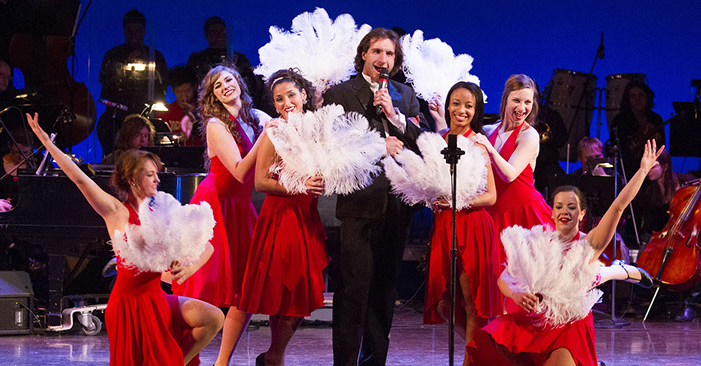 A man holds a microphone and sings, surrounded by dancers in red dresses and white feather fans during a fundraiser concert