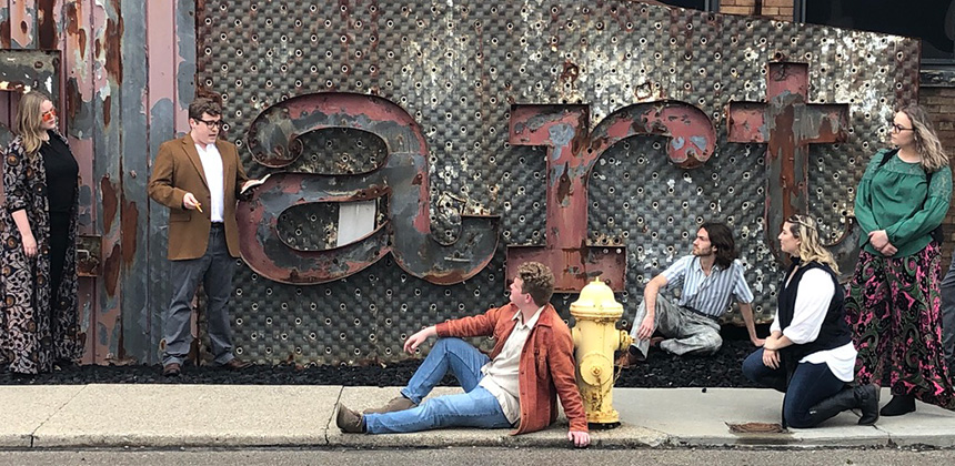 A small group of young people dressed in 60's era clothing talking together in front of an old rusty sign that reads "art"