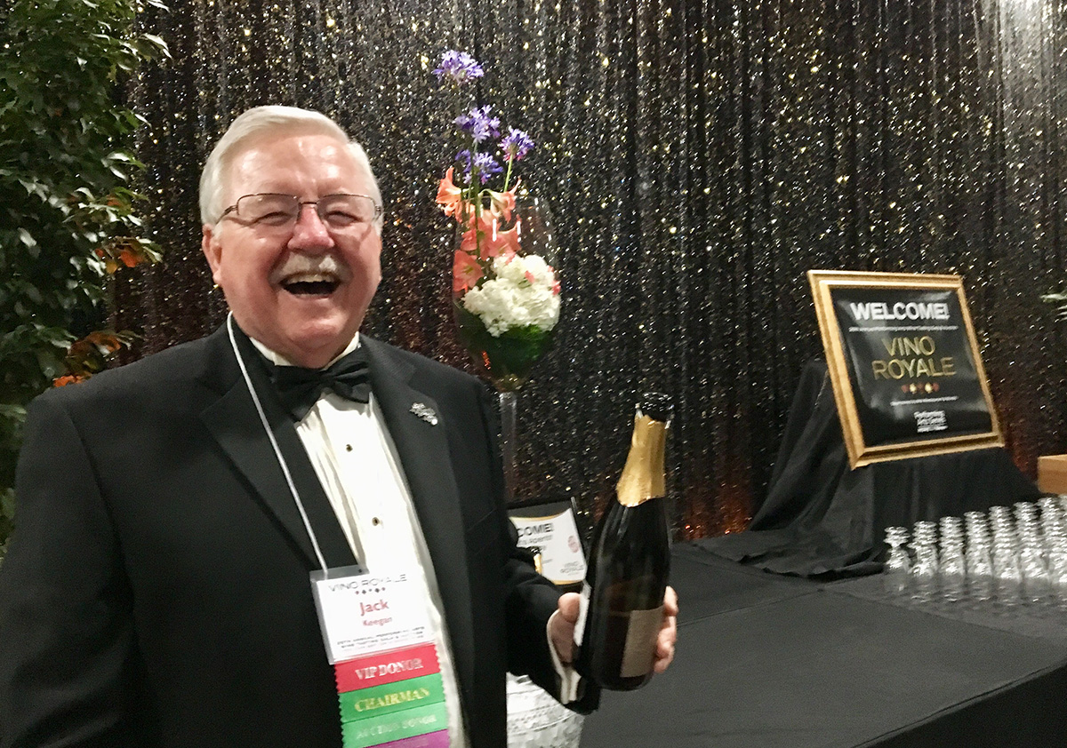 Jack Keegan smiles and holds up a bottle of wine near the welcome table at 2018 Wine Tasting