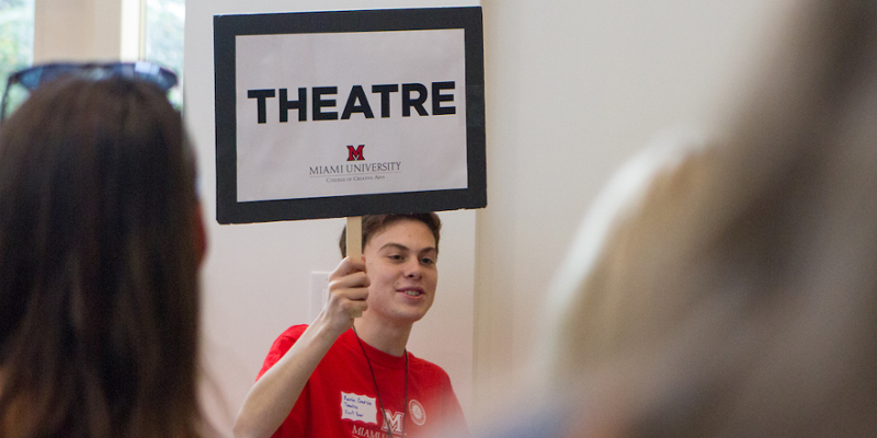A student holds up a sign to gather people who are interested in learning more about the Theatre program during an Arts Day event