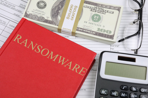 A picture of a red book with ransomware written on the cover together with a pile of 100 dollar bills, a calculator, a pair of glasses and papers.