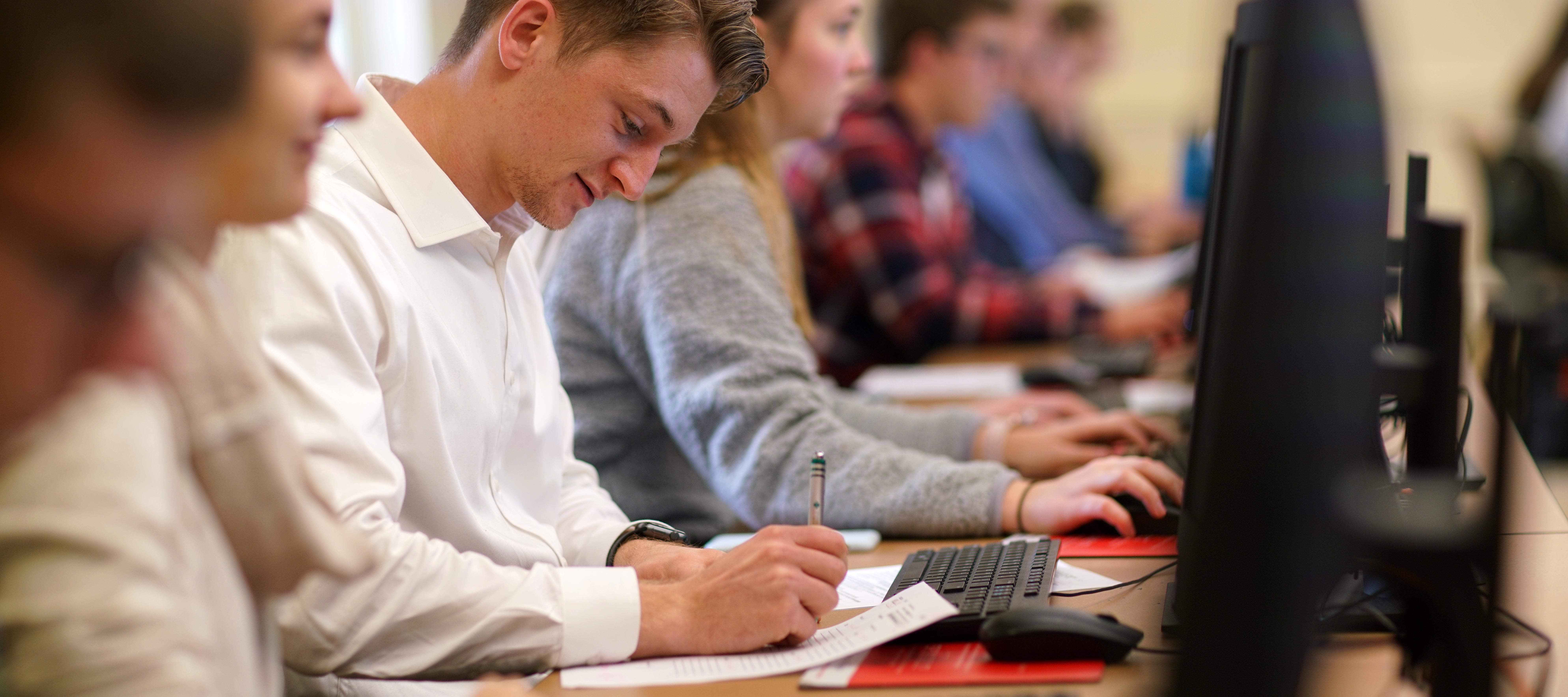 Male student writing on paper in front of computer in class