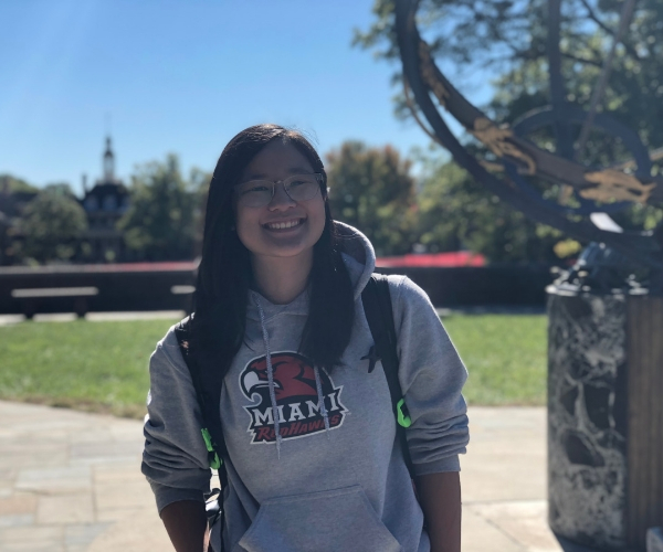 A miami student smiles in a Miami sweatshirt in front of MacCracken Hall