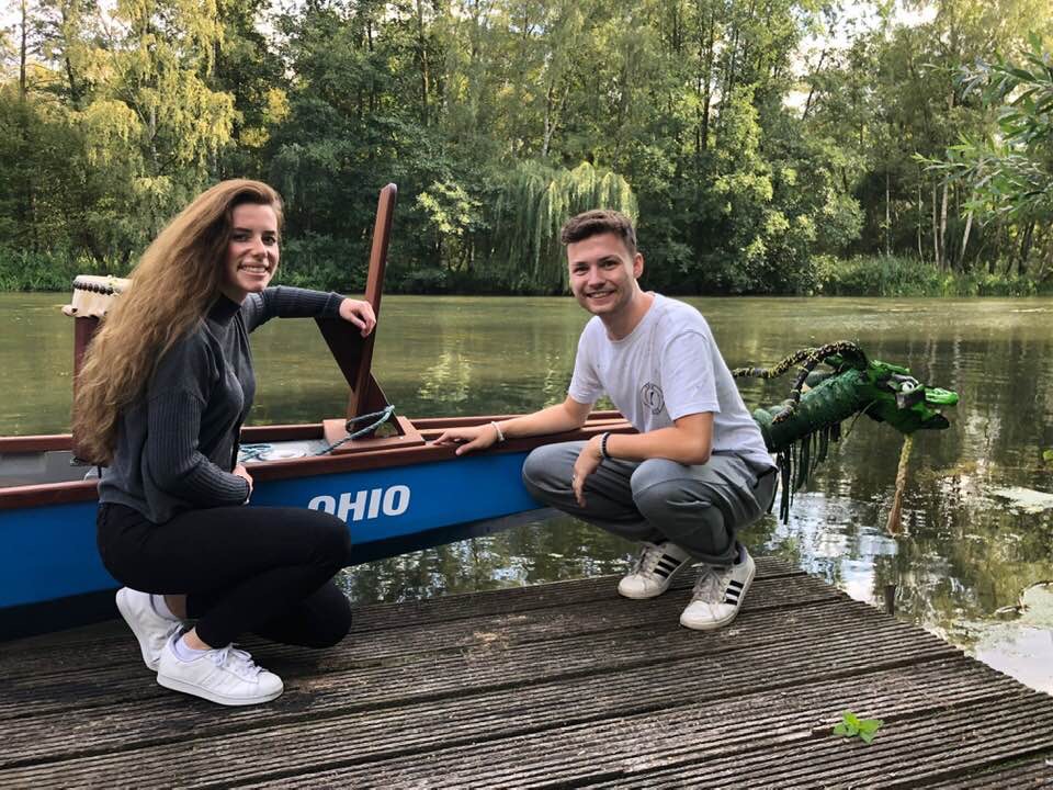 two students smiling in Germany kneeling in front of a small boat