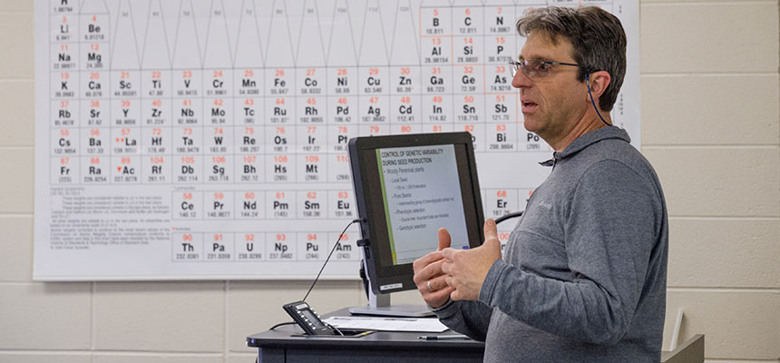 Brian Grubb standing in the front of the classroom teaching with a periodic table behind him.