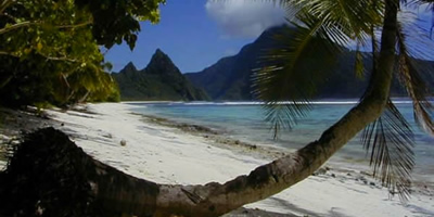 Mountains in distance, white sandy beach and turquoise sea in foreground, framed by tropical foliage.