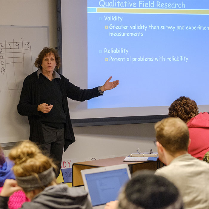 A professor teaching in the front of the class standing next to the projector screen.