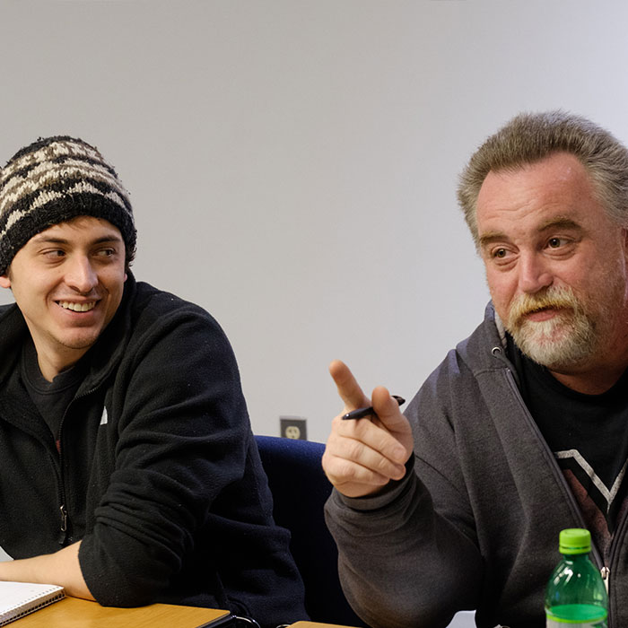 Two male students sitting at their desks talking to the class.