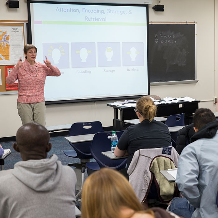 A female professor standing in the front of a classroom of students next to the projector screen.