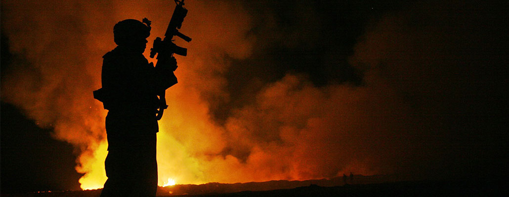 A soldier standing with in his gear with his gun pointed toward the sky with fire in the background.