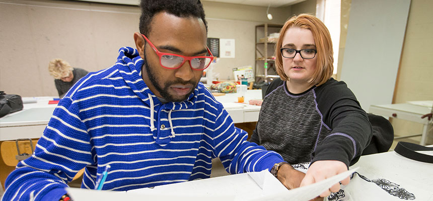A female and a male student sitting looking at a piece of paper during a printmaking class
