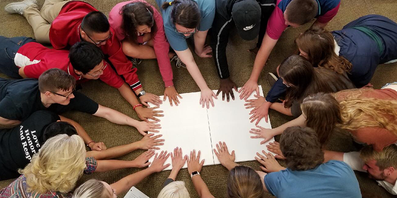 Students on the floor with one hand on a piece of paper all around the border.