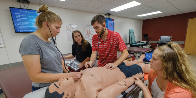 3 students and their professor using a stethoscope on a mannequin. 
