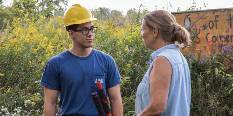 A student in a hard hat with hedge clippers in a field of all bushes