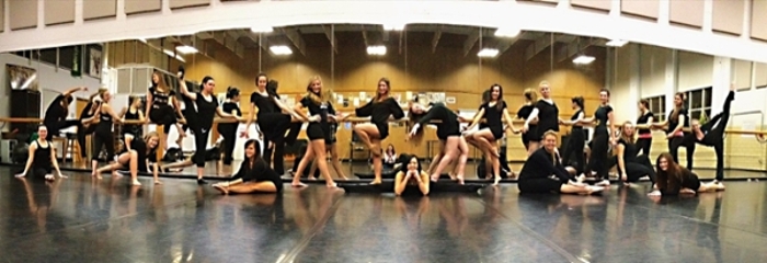Group of about 20 women in black leotards posing in various dance moves in front of wall-to-wall mirrors