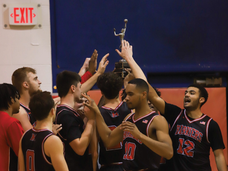 Harriers Men basketball team in a huddle holding up the MUM-OX-MUH trophy after beating Miami Middletown.