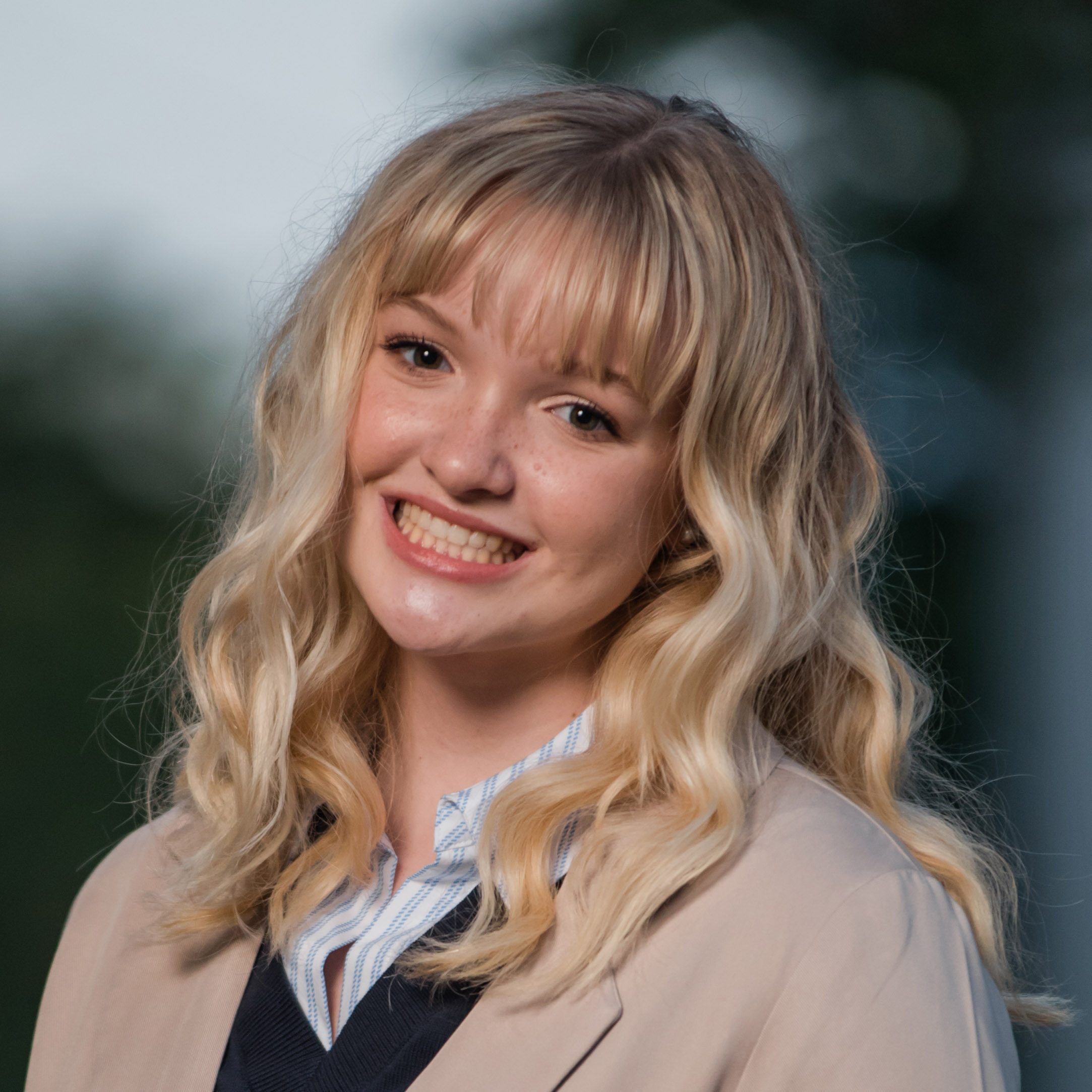 Head shot of first place winner Meredith Perkins. We see Meredith in the foreground smiling with long light blond hair and wearing a tan trench coat over a black sweater vest and a white and light blue pins-stripped shirt