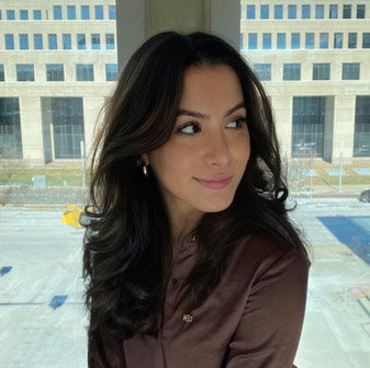 Sophia stands in the foreground wearing a brown button up shirt she is looking off to the right with a slight grin and her shoulder length brown wavy hair down. She stands at the intersection two large windows and we see a parking lot and a sandy brown stone building with many windows in the background.  