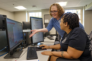 Two female-presenting people in front of a computer screen. One appears to be showing the other something on the computer.