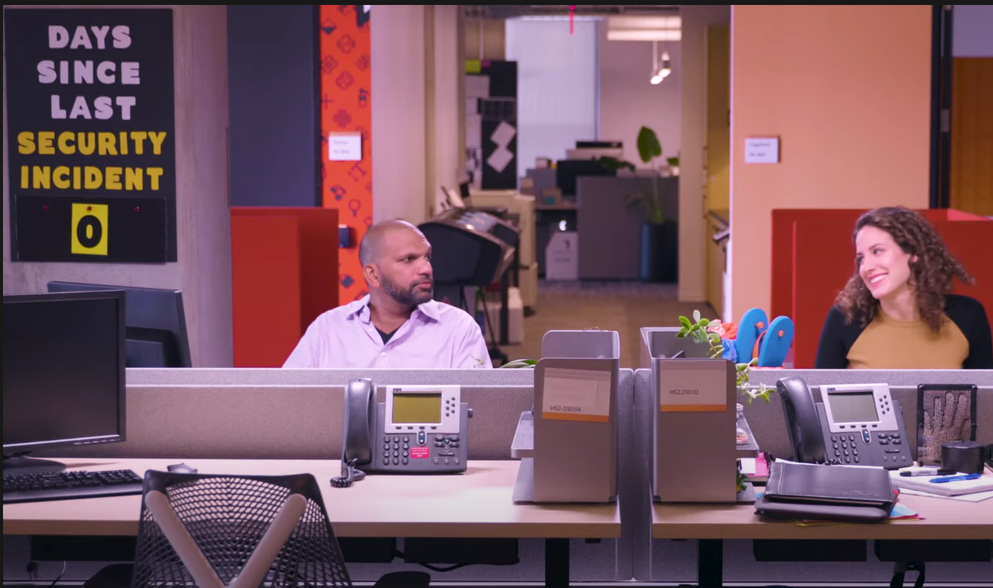 Two co-workers sitting in an office with a sign behind one that says zero days since last security incident