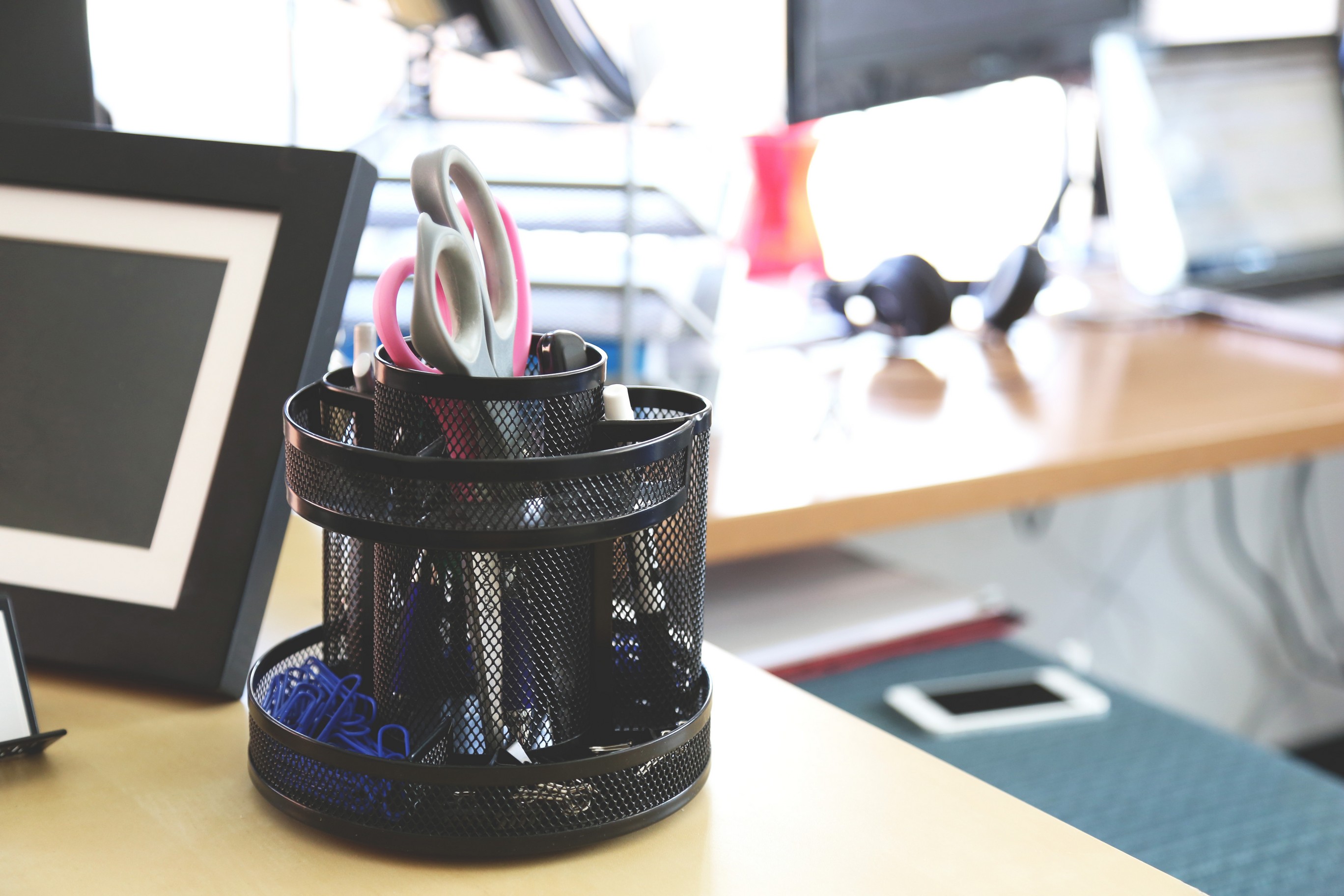 A round mesh container holding scissors, markers and paperclips is sitting on an office desk