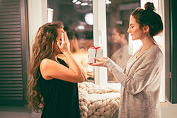 Two female-presenting people give each other gifts. One is holding out a box and the other is closing their eyes in anticipation