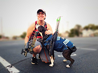 Naomi Cole kneeling with her service dog, Rhu. Rhu is wearing a blue service dog uniform and both are smiling