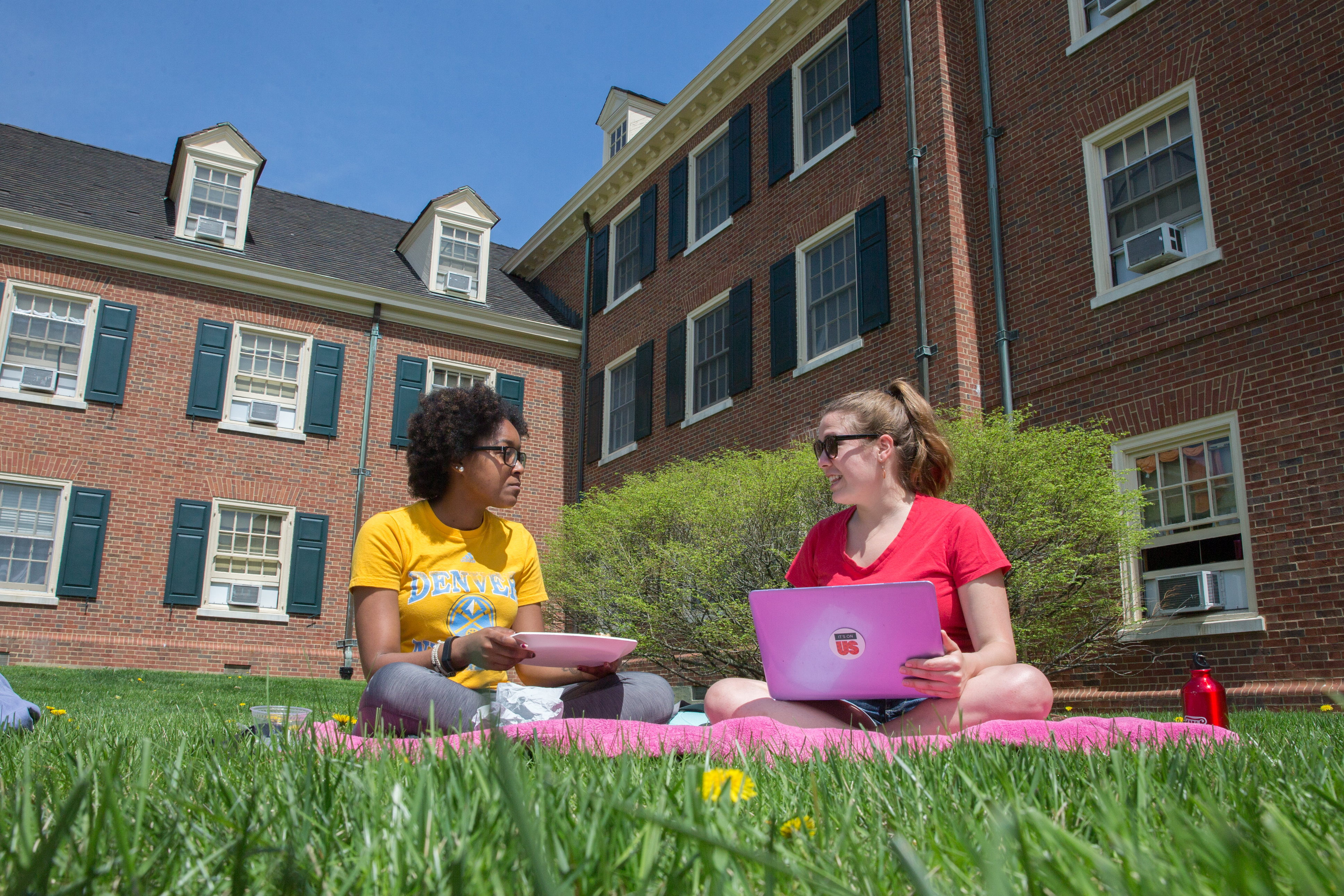 Two students sitting in the grass outside a dorm talking and working on their laptops