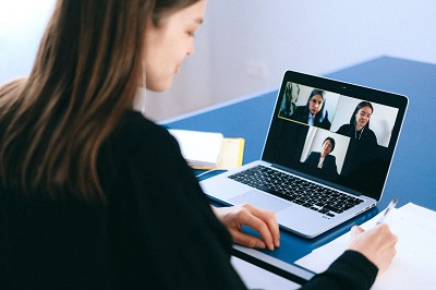 A female-presenting person writes notes on a notepad next to a laptop screen showing a video conference with three other people